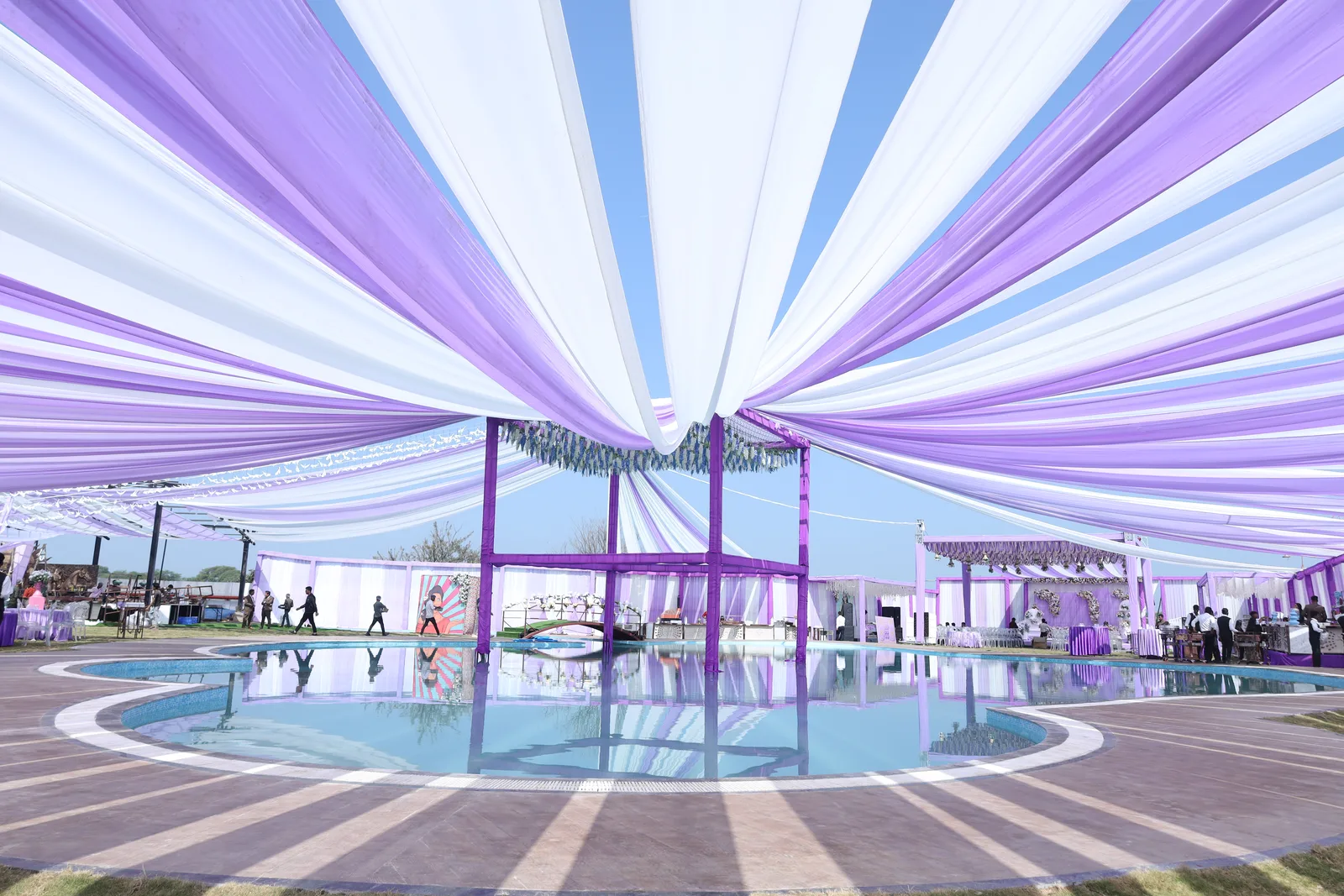 Still water and curved stone edge of the 6,500 sqft beach pool at Vetiver Grand, Jabalpur, violet canopies reflected across its surface.