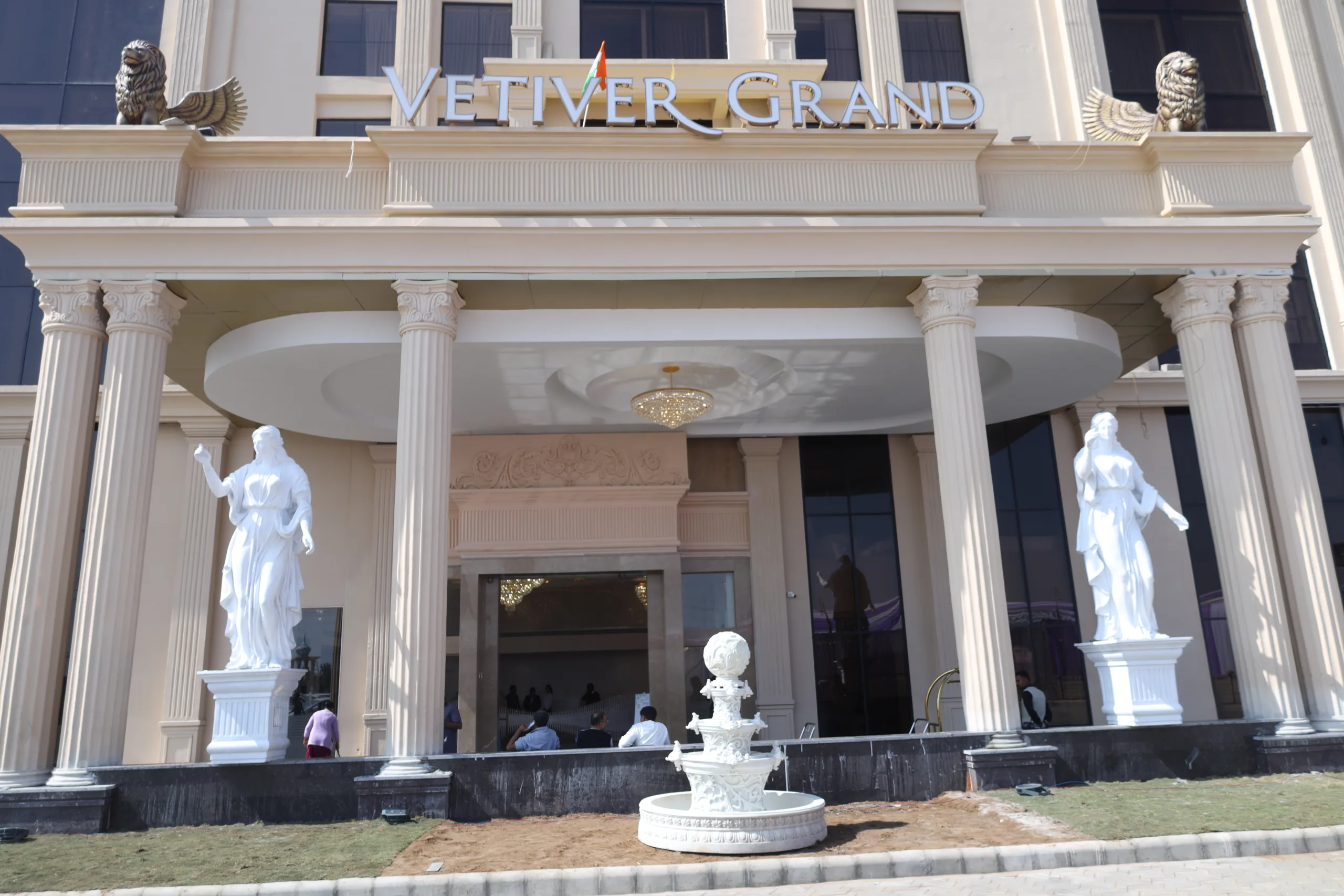 Daylight facade of Vetiver Grand, a luxury hotel in Jabalpur: hotel signage above the porte-cochère, flanked by golden winged-lion statues, classical columns and white goddess sculptures.